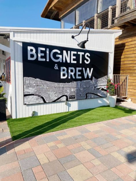 The outside of the Beignets & Brew coffee shop located on the Destin Harbor with its stark black and white contrasting sign and bright green fake grass.