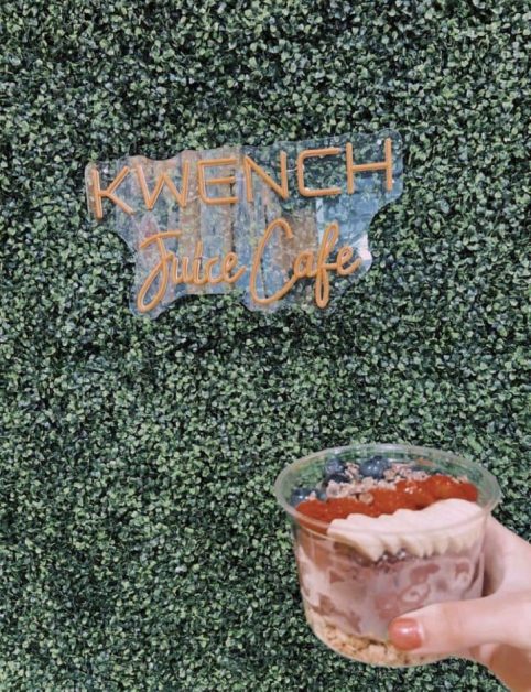 My hand holding the acai bowl I got in front of the greenery wall at Kwench Juice Cafe in Destin Florida.