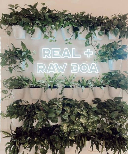 The greenery garland wall with a neon sign that reads Real + Raw 30A which is a cafe located along the 30A highway near Destin, Florida.