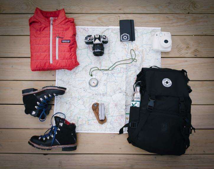 The hiking essentials laid on a map to prepare for an upcoming camping trip consisting of a travel backpack, water bottle, hiking boots, compass, and a red Patagonia wind breaker.