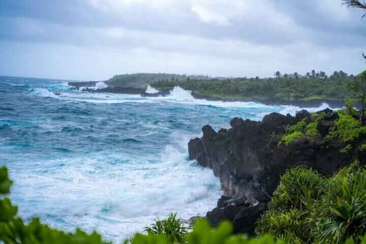 An overlook view to the powerful blue ocean water, black rugged coastline, and dense green vegetation at Wai'anapanapa State Park in Maui.