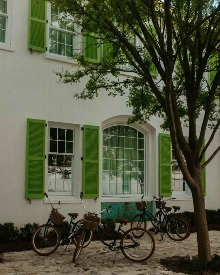 A collection of bikes with baskets on the front placed in front of a white clay beach house with lime green shutters. Biking is one of the best things to do in 30A, Florida.
