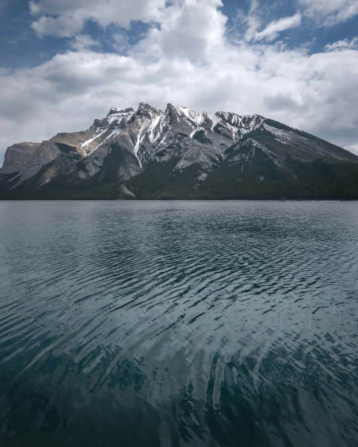 A gloomy and overcast day at Lake Minnewanka in Banff National Park with its dark blue waters surrounded by snow covered mountains and dark grey rain clouds.