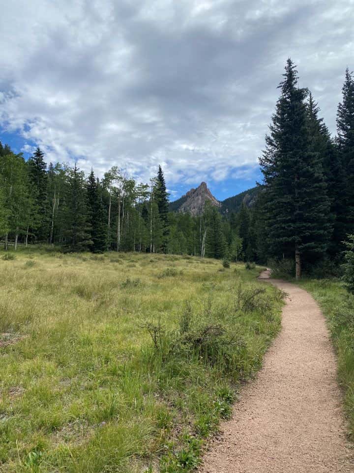 The gravel sand-like path you follow along the Crag's Trail surrounded by tall pine trees, grassy areas, and rock formations and mountains in the distance.