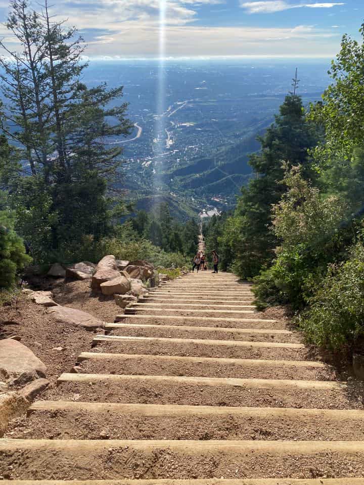 The steep sandy stairs that you climb to reach the top of the Manitou Incline are surrounded by green trees, rocks, and views of the surrounding area.