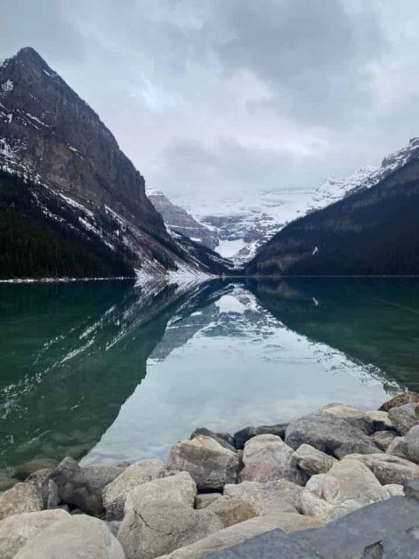 The clear glassy reflections of Lake Louise surrounded by the stone rocks on the shore and mountain range in the distance.