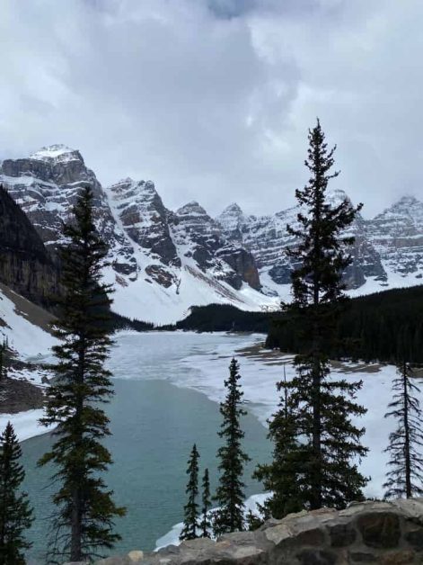 The views of the partially frozen Moraine Lake surrounded by snow covered mountains and dark green fir trees in late May 2024.