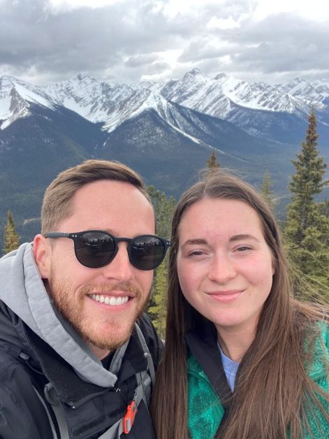 My husband and I taking a photo of ourselves with the stunning Canadian Rockies mountain range in the distance that are still partially snow covered in late May which you can see from the Banff Gondola summit.