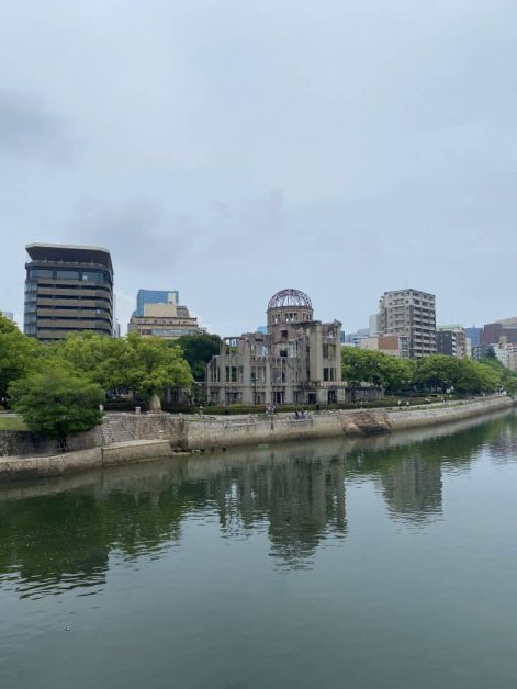 The Atomic Bomb Dome in Hiroshima, Japan in June, with surrounding modern buildings and trees reflecting in the Motoyasu River on a cloudy day