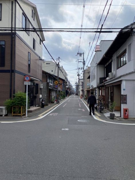 Quiet back street in Kyoto lined with traditional and modern buildings, power lines overhead, and a person walking down the middle of the empty road on a cloudy day.