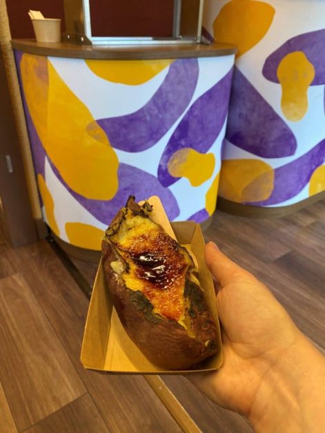 A hand holding a crème brûlée sweet potato from Imo Pippi in Kyoto, with a colorful purple and yellow counter in the background.