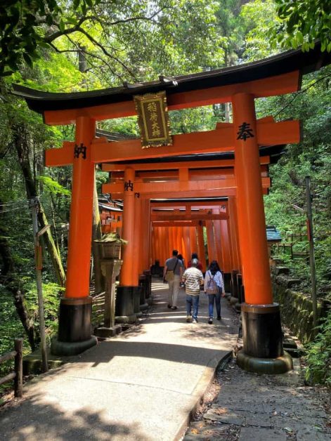 People walking through the thousands of bright red/orange torii gates lining the path to Fushimi Inari Taisha shrine in Kyoto.
