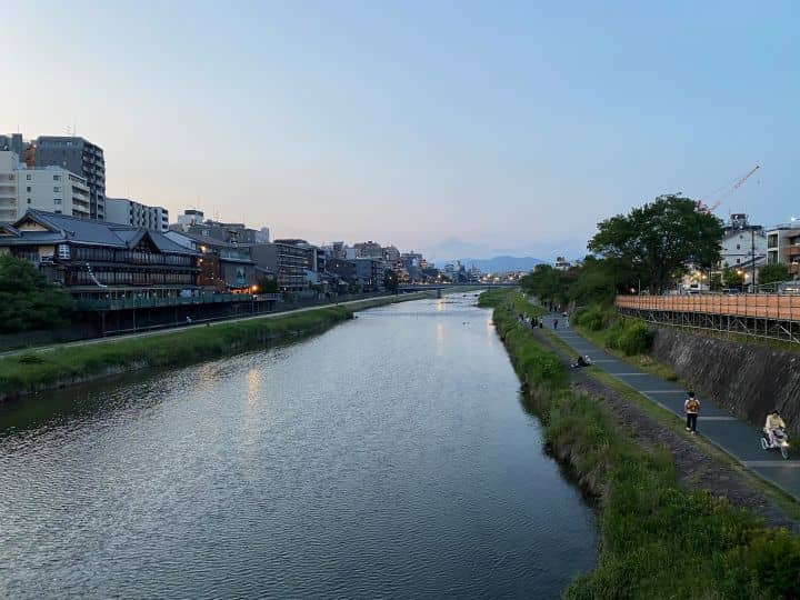 People walking on the paths running along the Kamo River in Kyoto at sunset with the glow of the sun emitting orange colors in the sky.