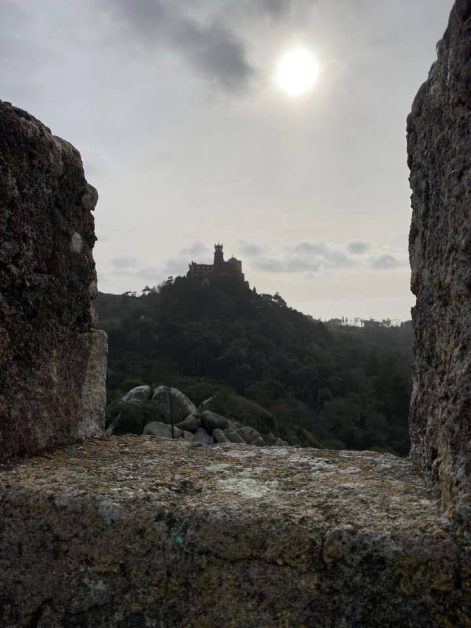 The sun shines through a stone wall window, highlighting Pena Palace's beauty from the Moorish Castle in Sintra.