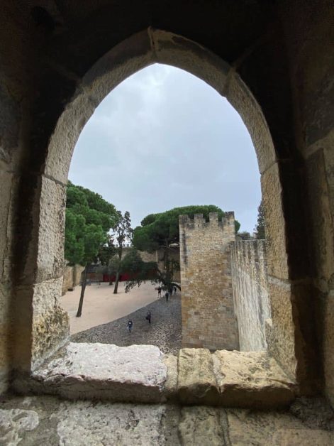 A view through a stone arched window which shows the courtyard with cobblestones and lush green trees at Castelo de Sao Jorge in Lisbon.