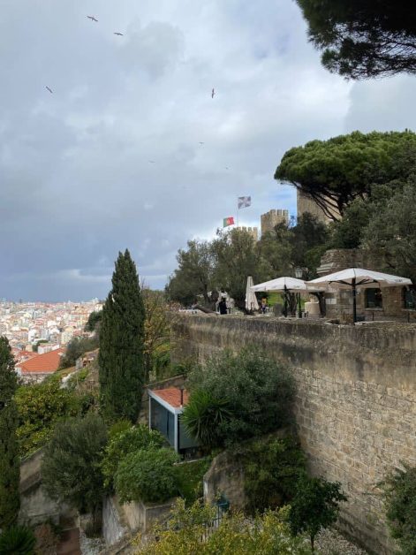 Castelo de Sao Jorge offering a view of a historic stone wall with umbrellas and people, overlooking a cityscape under a cloudy sky.