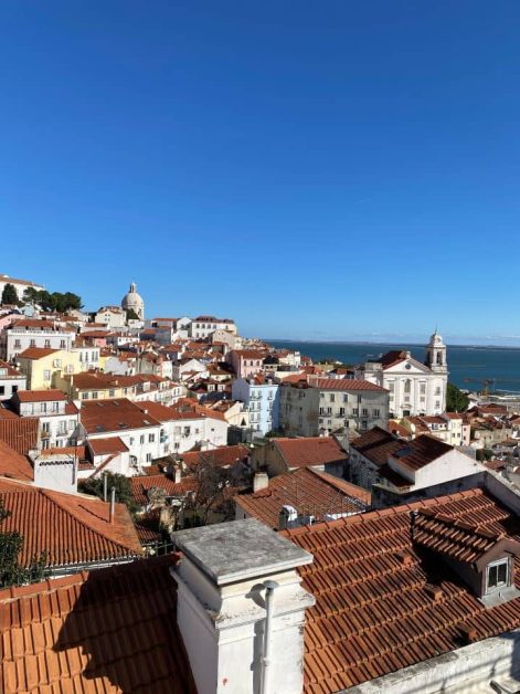 A scenic view of Lisbon from Miradouro das Portas do Sol with tightly packed terracotta-roofed buildings under a clear blue sky. The ocean is visible in the background,