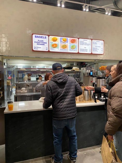 Inside Apollo Bagels in New York City with customers ordering at the counter and menu board displaying bagel options.