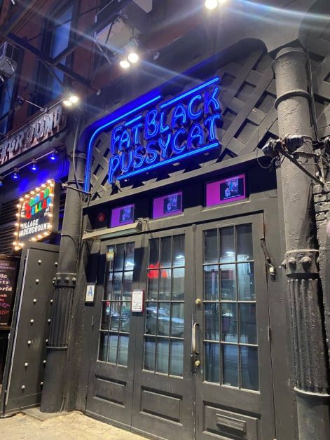 The dark black exterior and blue neon sign outside the Fat Black Pussycat which is one of the Comedy Cellar locations in New York City.