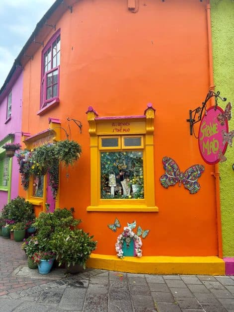 Colorful street corner in Kinsale with a bright orange building housing "Stone Mad" jewelry shop, yellow-trimmed windows, decorative butterfly ornaments, and potted plants on the sidewalk.