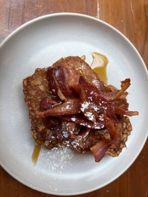 Overhead view of a plate of French toast topped with crispy bacon strips, maple syrup, and a dusting of powdered sugar, on a wooden table.