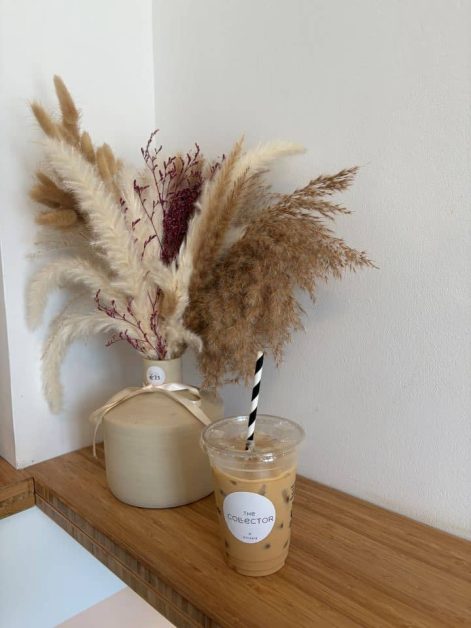 An iced latte in a clear cup with a black-and-white striped straw, labeled "The Collector," sitting on a wooden shelf next to a vase of dried pampas grass.