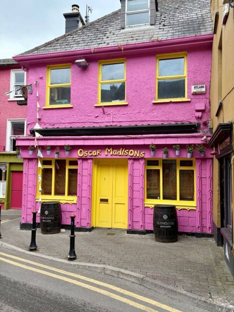 Bright pink building facade with yellow trim, reading "Oscar Madisons" above a yellow door, flanked by cider barrels on the street.