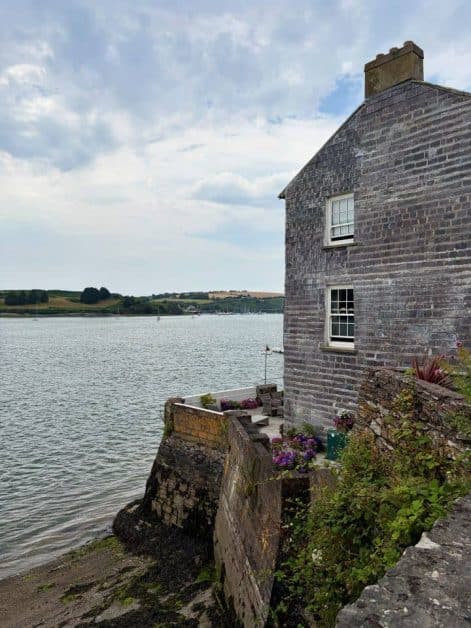 A stone cottage perched at the water's edge with a small terrace and flowering plants, overlooking a harbor with distant green hills.