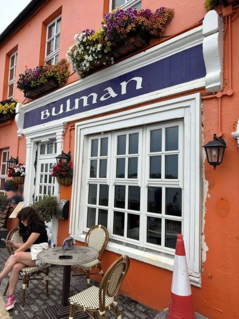 Orange building exterior with a navy blue sign reading "Bulman" in white lettering, flower boxes above the windows, and outdoor café seating where a woman sits at a table.