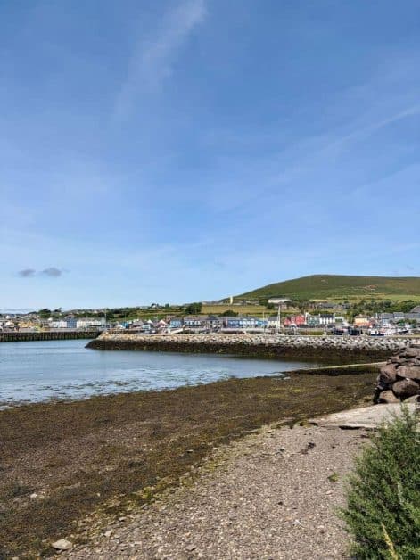 View across Dingle Harbor's low-tide shoreline toward the colorful buildings of town and a green hillside beyond.