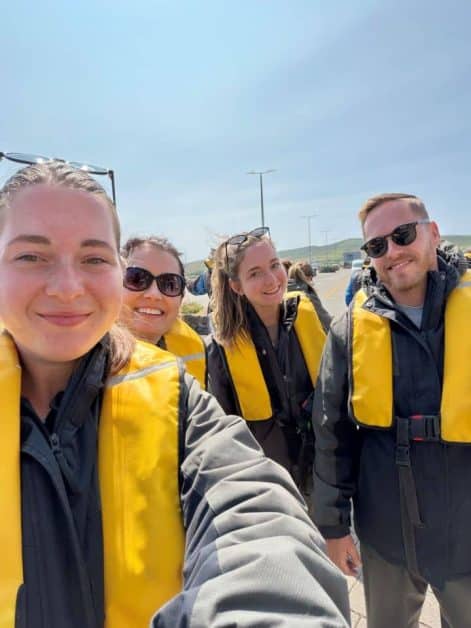 Selfie of four smiling people wearing yellow life jackets, preparing to board a boat with hills visible in the background.