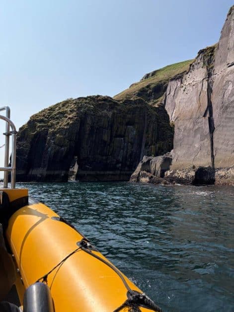 View from a yellow inflatable boat approaching tall layered cliff formations rising from the sea.