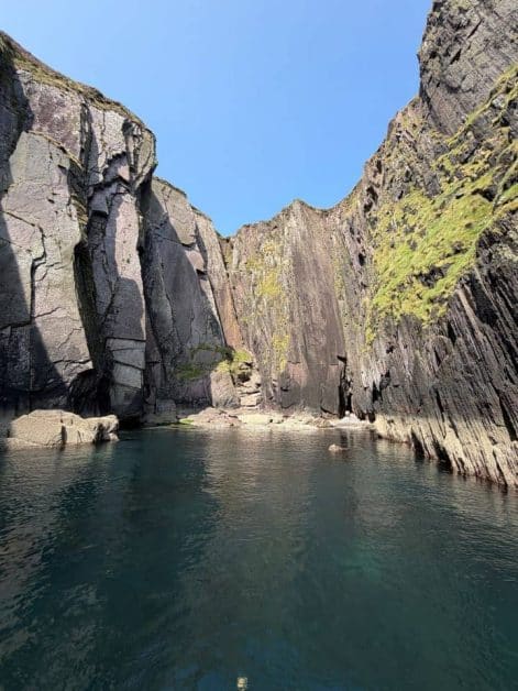 Dramatic rock walls flanking a narrow sea inlet with calm turquoise water, viewed from a boat.