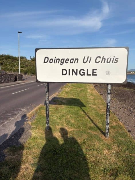 Bilingual road sign reading "Daingean Uí Chúis / Dingle" on a grassy roadside strip, with two people's shadows cast in front of it.