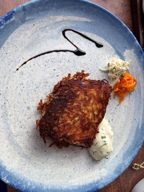 Overhead view of a crispy potato cake plated with herb cream sauce, shredded carrot and cabbage slaw, and a balsamic drizzle.