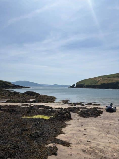 Secluded sandy beach with seaweed-covered rocks, a small inflatable boat, and views of a rocky headland and distant mountains across the water.