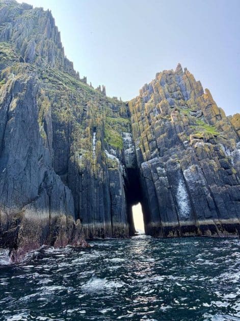 Tall jagged sea cliffs with a narrow natural archway opening to light beyond, viewed from the water.