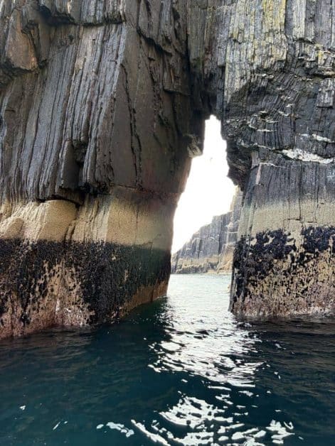 Close-up of a layered rock archway with mussel-covered base, framing a glimpse of open sea and cliffs beyond.