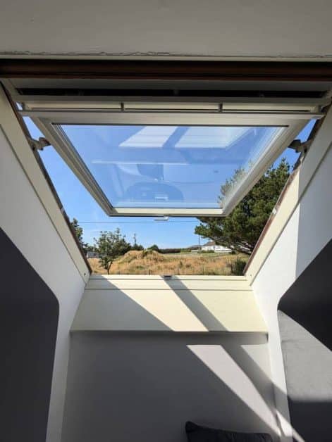 Open skylight window in an attic room showing blue sky, trees, and a countryside view.