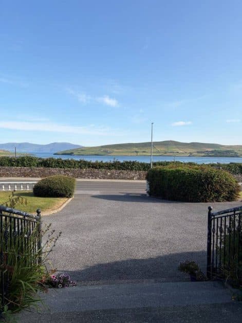 View from a driveway through wrought iron gates toward a coastal road, bay, and distant green hills under clear skies.