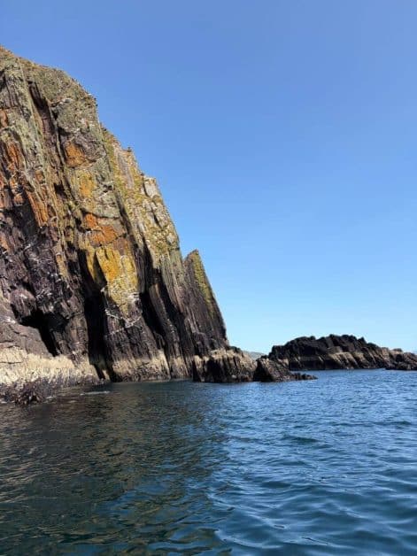 Sunlit layered cliffs with orange and green lichen streaks rising from the sea, with smaller rock formations visible offshore.
