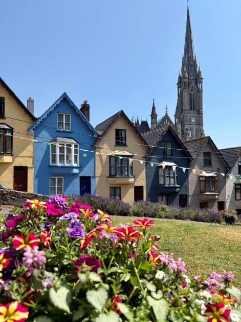 Colorful hillside houses in blue, yellow, and gray with Cobh Cathedral spire behind, bright flowers blooming in the foreground.