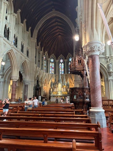 Interior of Cobh Cathedral with high vaulted ceilings, rows of wooden pews, stone columns, an ornate carved pulpit, and stained-glass windows above the altar.