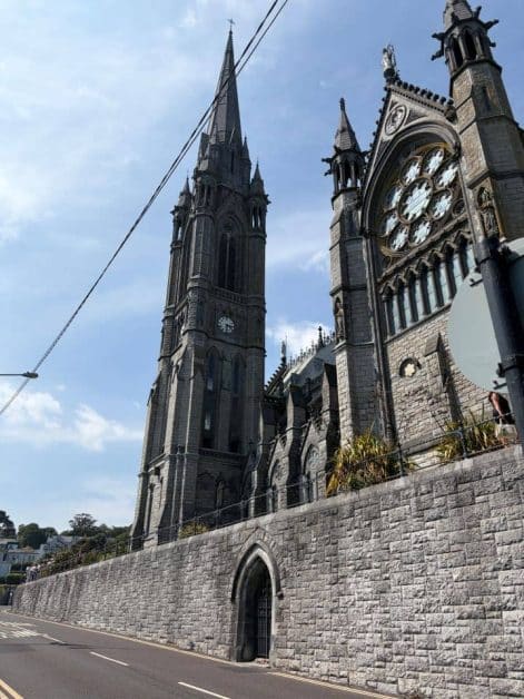 Exterior view of St. Colman's Cathedral's tall Gothic spire and rose window, seen from the street beside a stone retaining wall.